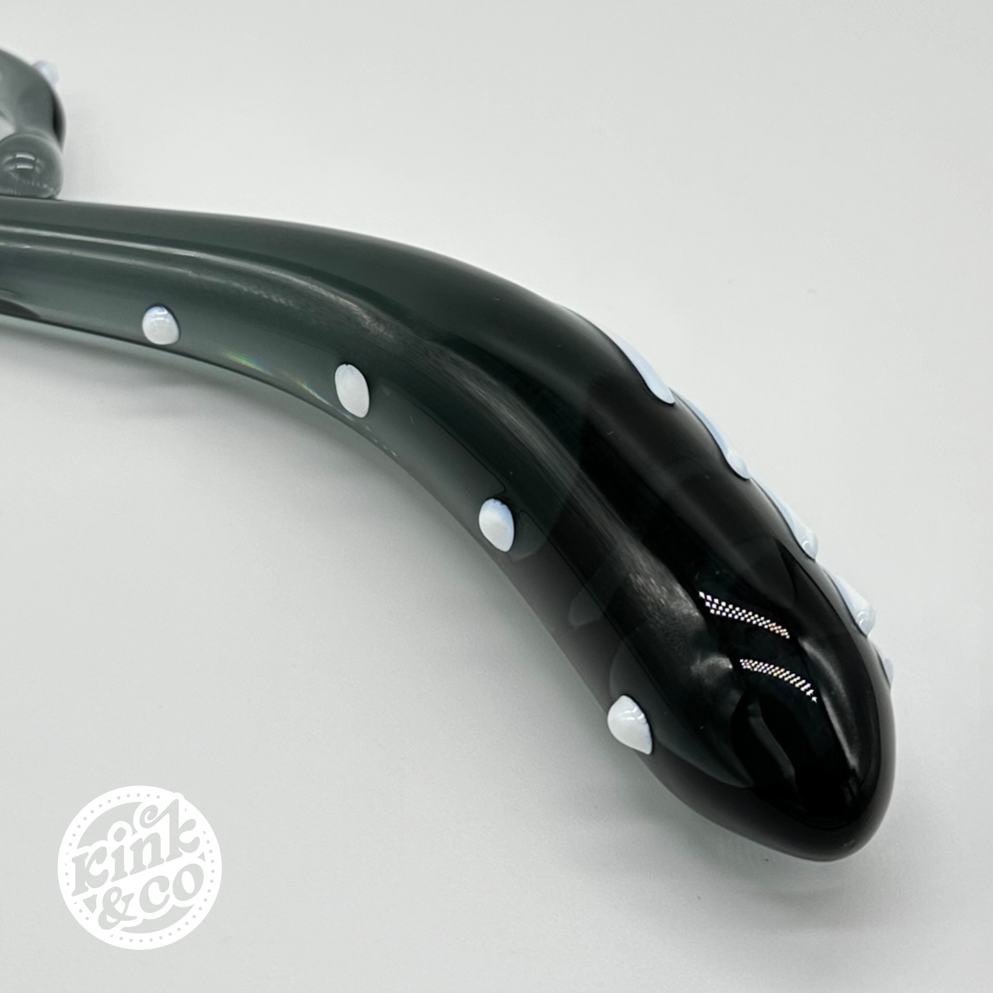 Close up of the thick end of a black and white sex toy made from black glass. Grey background with the kink and co logo in the bottom left of the image 