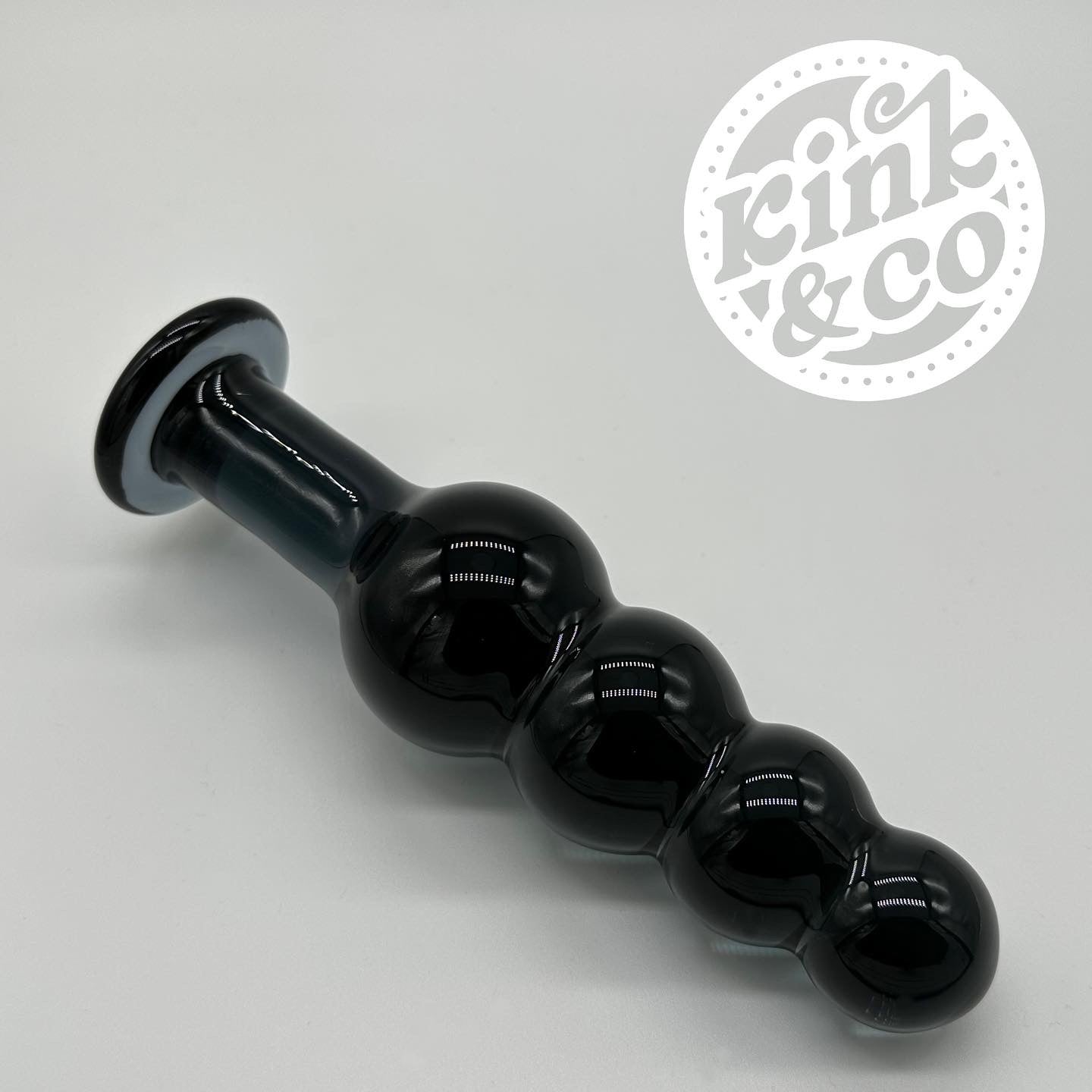 Beaded sensual black glass dildo laid on its side. A grey background with the kink&Co logo in the top right hand side of the image 